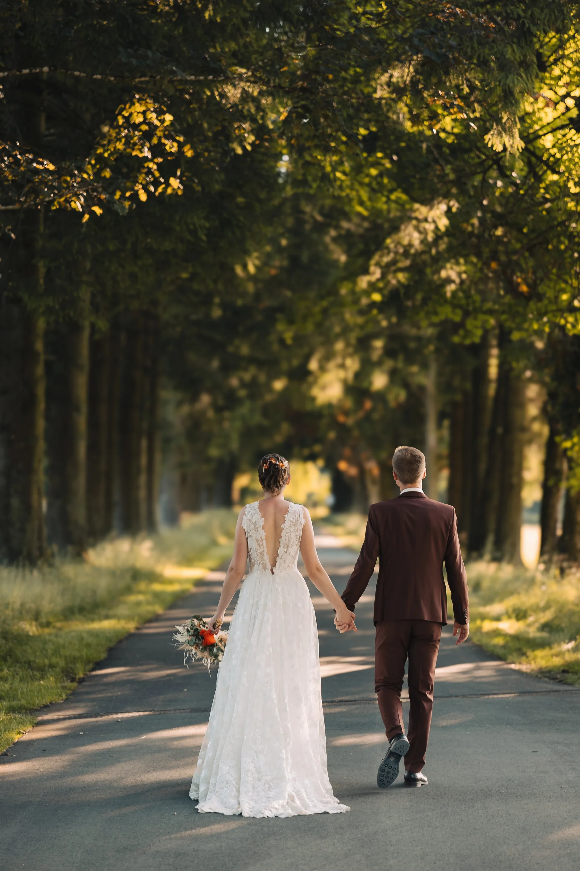 Les mariés de dos, main dans la main, sous une allée d'arbres