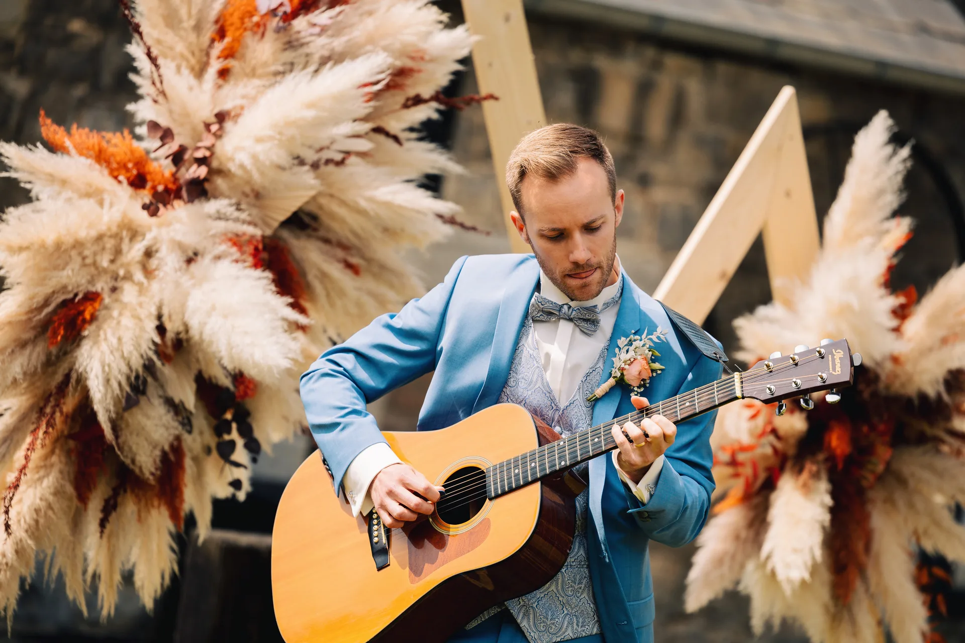 Le marié joue de la guitare acoustique devant une arche de fleurs séchées